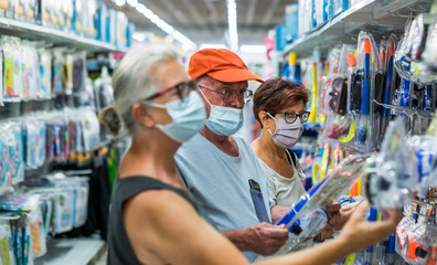 Fototapeta na wymiar Three adult senior people shopping together in sea accessories department choosing a scuba mask, wearing medical mask due to coronavirus - new normal concept and retired elderly