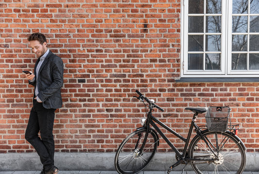 Young Businessman Going To Work On Bike Commute Using His Mobile Cellphone Against City Brick Wall Background. Happy Business Man Bicycle Commuter Arriving At Office Using Phone Texting.