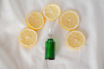 Green bottle of lemon essential oil with lemons slice on white fabric background. Top view flat lay. Beauty cosmetic natural skincare product mock up.