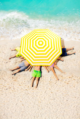 Group of kids lay on the beach sand under big sun umbrella view from above near sea waves