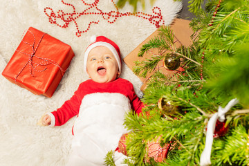Happy little toddler boy lay on the carpet with open mouth laughing and looking at camera under Christmas tree
