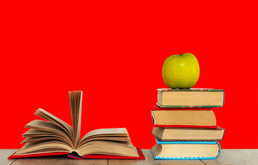 Open book textbook, pile of books and green apple on a wooden desk on the red background. Back to school distance home education.Quarantine concept of stay home.