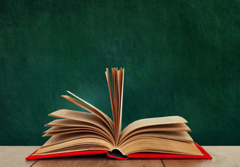 Open book textbook in a red cover on a wooden desk on the background of a green school chalkboard. Back to school distance home education.Quarantine concept of stay home.