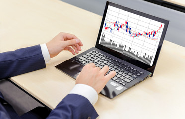 ノートパソコンでモニター上の株価チャートを見るビジネスマン