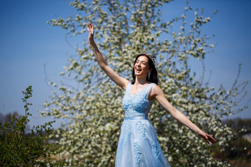 Young woman with beautiful make-up with a vintage hoop in a blue light dress with a bouquet posing on a background of green foliage in a park. Fresh portrait attractive girl with white flowers.