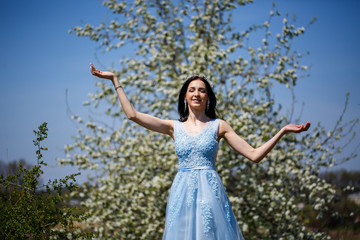 Portrait of a tender girl in a blue long dress with tulle under blooming cherry with a smile on her face on a sunny warm summer day