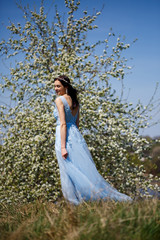 Fototapeta premium Portrait of a tender girl in a blue long dress with tulle under blooming cherry with a smile on her face on a sunny warm summer day