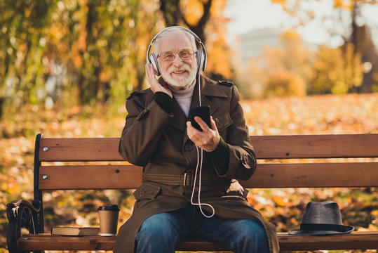 Photo of senior retired old grey haired grandpa feel young street sit bench listen cool youth song big earflaps close to grandchildren cheerful positive emotions autumn jacket outdoors