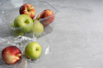 Plastic or polyethylene transparent bag and container full of ripe apples and nectarines at kitchen on concrete background showing environmental pollution problem. Image with copy space