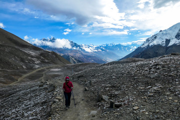 A woman in pink jacket walking on a pathway leading to Tilicho Lake, Nepal. Stormy weather in snow capped Himalayan peaks along Annapurna Circuit. Barren and sharp slopes. Coldness