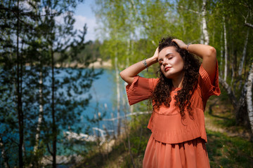 Naklejka premium A girl with long wavy curly hair in her hair woven flowers in a white transparent guipure dress barefoot in the summer on nature in a forest at sunset standing standing near the road and bushes