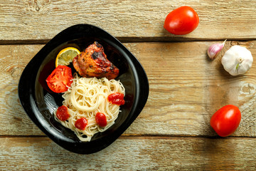 Black plate of spaghetti with baked chicken on a wooden table near vegetables.