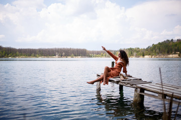 Fototapeta na wymiar Young woman with long wavy curly hair in a long guipure dress barefoot in the summer in a forest on a lake at sunset standing on a pantone on a wooden pier bridge. Summer sunny day