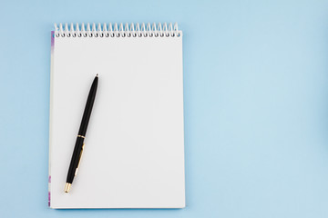 Blank white notebook with a black branded pen on a light blue background. View from above. Education concept.