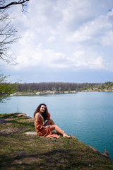 A young woman with curly hair and a smile on her face sits in a green meadow on a background of blue lake. Warm summer day, happy girl, emotions of joy