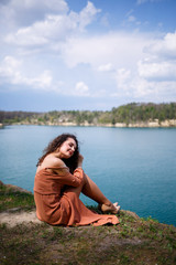 A beautiful girl of European appearance with curly hair and a smile on her face sits in a green meadow against the background of a blue lake. Warm summer day, happy young woman, emotions of joy