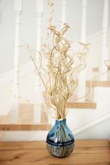 Floral composition of dried branches in blue vase on wood table with white wood staircase on background, copy space. Home interior decor