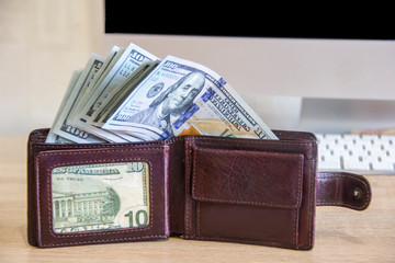 Packs of money are in a wallet that lies on the table near the computer. Money dollars lie in a leather wallet that stands on the table in the office.