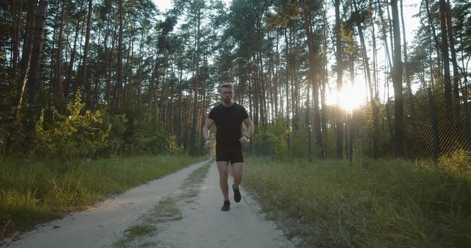 Young active guy in black t-shirt, shorts and sneakers running on road among summer forest. Sporty man with muscular body enjoying beautiful nature during workout.