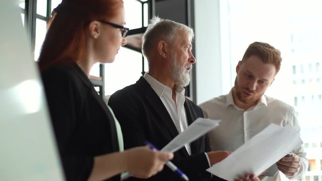 Three business people examining financial data on tablet computer standing near big window background of cityscape. Business team brainstorming in modern executive office. Tracking shot ins low motion