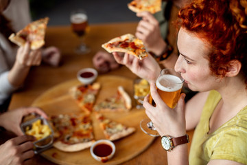 Redhead woman drinking beer while eating pizza with friends in a pub.