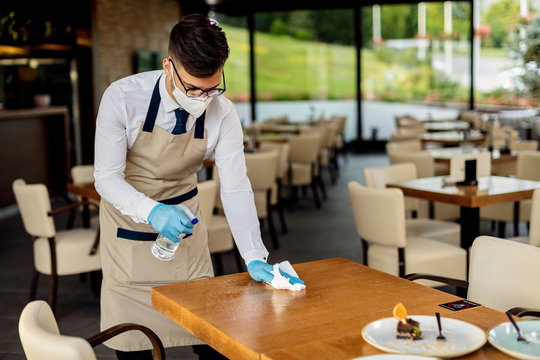 Waiter with protective face mask disinfecting tables in a cafe due to coronavirus epidemic.