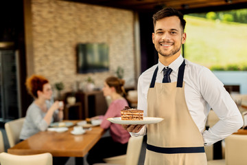 Young happy waiter serving chocolate dessert to customers in a cafe.