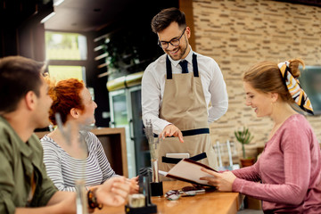 Young happy waiter communicating with group of customers in a cafe.
