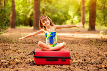 Little child girl wear swimsuit sitting on suitcase in summer forest with hands up, show plane. Vacation concept