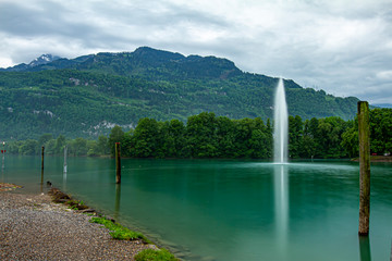 Landscape photo of a mountain lake amongst Swiss Alps. The long exposure photo features a water jet fountain in the  lake ejecting high pressure water into the air. A forest surrounds lake.