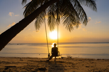 The guy enjoys the sunset riding on a swing on the ptropical beach. Silhouettes of a guy on a swing hanging on a palm tree, watching the sunset in the water.