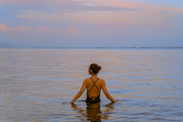 A beautiful girl bathes in the warm sea at sunset. The calm sea reflects the sunset colors of the sky. Tourist destination Bali, Indonesia. 