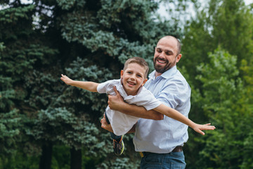 a bearded man rolls on the shoulders of a teenage boy in a park. Dad and son play airplanes in nature,