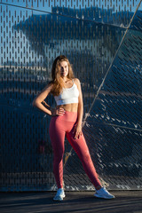 Portrait of sports woman in pink leggings and long hair posing against metal wall background, sunset light, outdoors. 