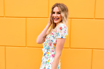 Bright sunny portrait of pretty blonde woman wearing floral sexy dress, posing near yellow wall, making surprising face, toned colors.