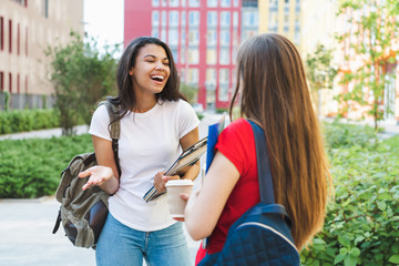 Two happy female students standing outdoors at the college campus smiling and talking to each other