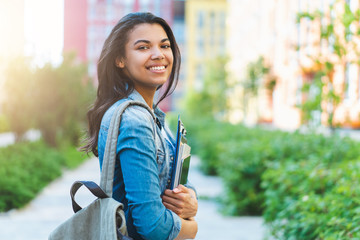 Half-length portrait of beautiful casually dressed student girl with charming smile posing outdoors with a backpack and pile of books and tablet in hands - 371322914