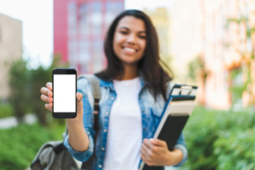Smiling african american girl student holding smartphone in hands demonstrating its white blank screen to the camera. Focus is on hand with phone. Mock up, copy space for your text or interface