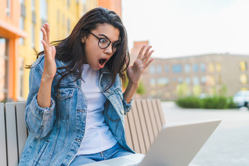 Young dark skinned woman blogger in trendy spectacles sitting on a bench outdoors and looking at her laptop screen with shocked open mouth face expression unable to believe  she got million followers