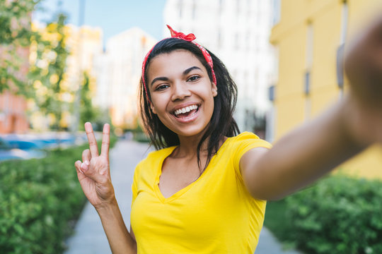 Portrait of beautiful casually dressed african american girl with perfect smile posing outdoors taking selfie and shoving peace sign