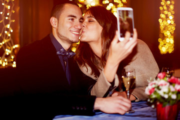 Portrait of a happy romantic couple making selfie photo with smartphone, cheerful girlfriend kissing her boyfriend on the cheek while taking self portrait