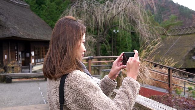 Happy young Asian woman tourist using smartphone taking a photo of old village at Yamanashi, Japan with beautiful nature in autumn. Beautiful lady relax and enjoy with autumn vacation travel in Japan