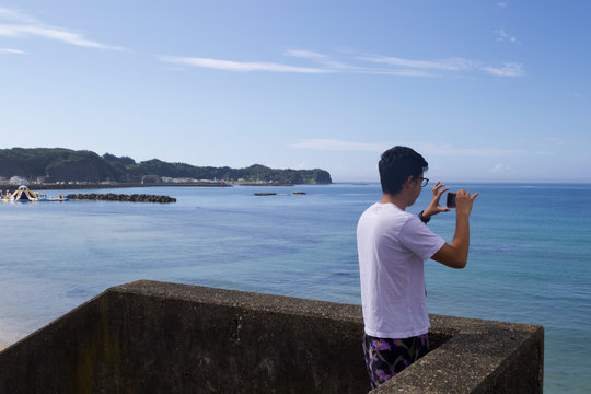 An Asian boy is taking pictures of a beautiful blue bay in Japan with his mobile device, it is a pretty scene with a wonderful landscape. Katsuura in Chiba is known for its many bays.