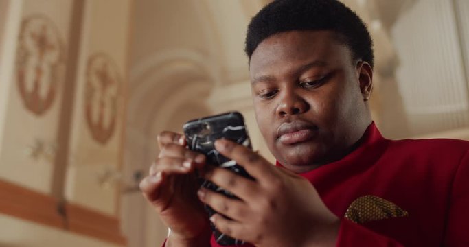 Close up view of african american man in red suit using his smartphone. Young male person looking and touching phone screen while searching gospel song words. Indoors