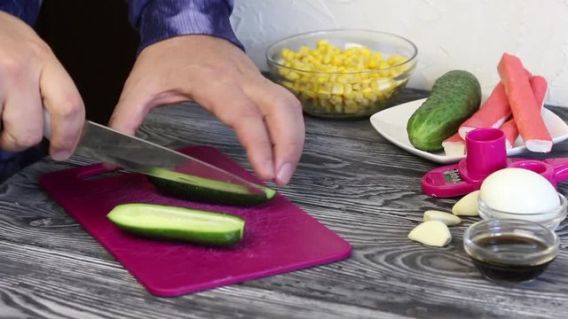 A man cuts a cucumber on a cutting board. Crab sticks, chicken eggs and vegetables for salad preparation. They lie on painted pine boards.