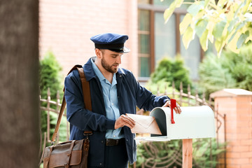 Handsome young postman putting letter in mail box outdoors
