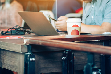 Man work on computer with smartphone and relax with hot coffee in coffee shop, where seats are spaced for social distancing during the Corona virus (COVID-19) epidemic. Selective focus on coffee cup.