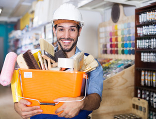 portrait of smiling foreman standing near racks in paint store holding basket with tools