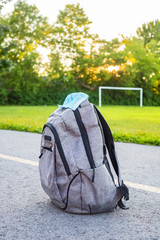 School backpack with medical mask on the ground, green soccer field and sunset background. Back to school during epidemic concept.