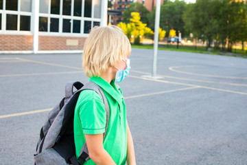 Kid in mask during corona virus outbreak. Little schoolboy wears grey backpack, breathes through mask, going to school. Back to school concept after reopening. Student at the school yard